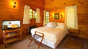 A cozy wooden cabin bedroom with a bed, a TV on a wooden stand, and a window with curtains. - Cabañas Playa Linda (Villarrica)