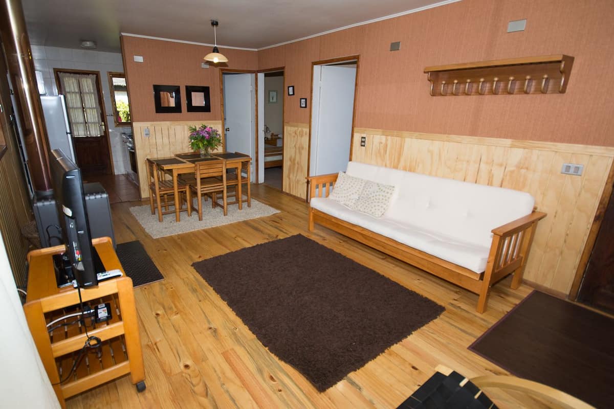 Un salon chaleureux avec un plancher en bois, un canapé blanc, une table à manger avec des chaises et un téléviseur posé sur un meuble en bois.
