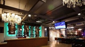Una zona de bar con mostrador, estantes llenos de botellas, una lámpara de araña y una pantalla de televisión.