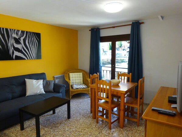 A living room with a dark sofa, a small table, a wicker chair, a dining table with chairs, a TV, and a window with curtains. - Apartamentos Arinsal 3000 (Arinsal)