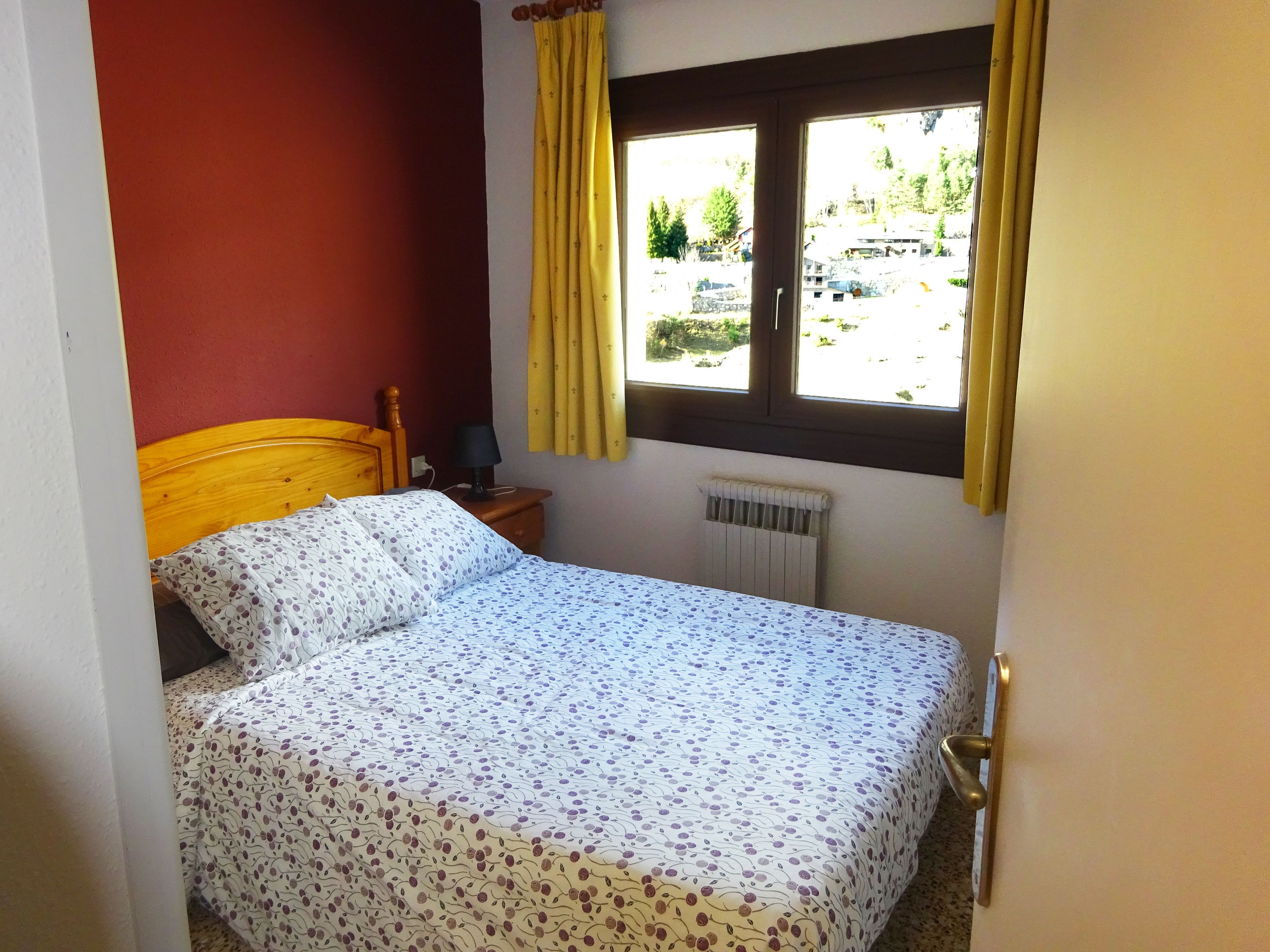 Un dormitorio con cama de madera, una mesita de noche con lámpara, una ventana con cortinas y un radiador.