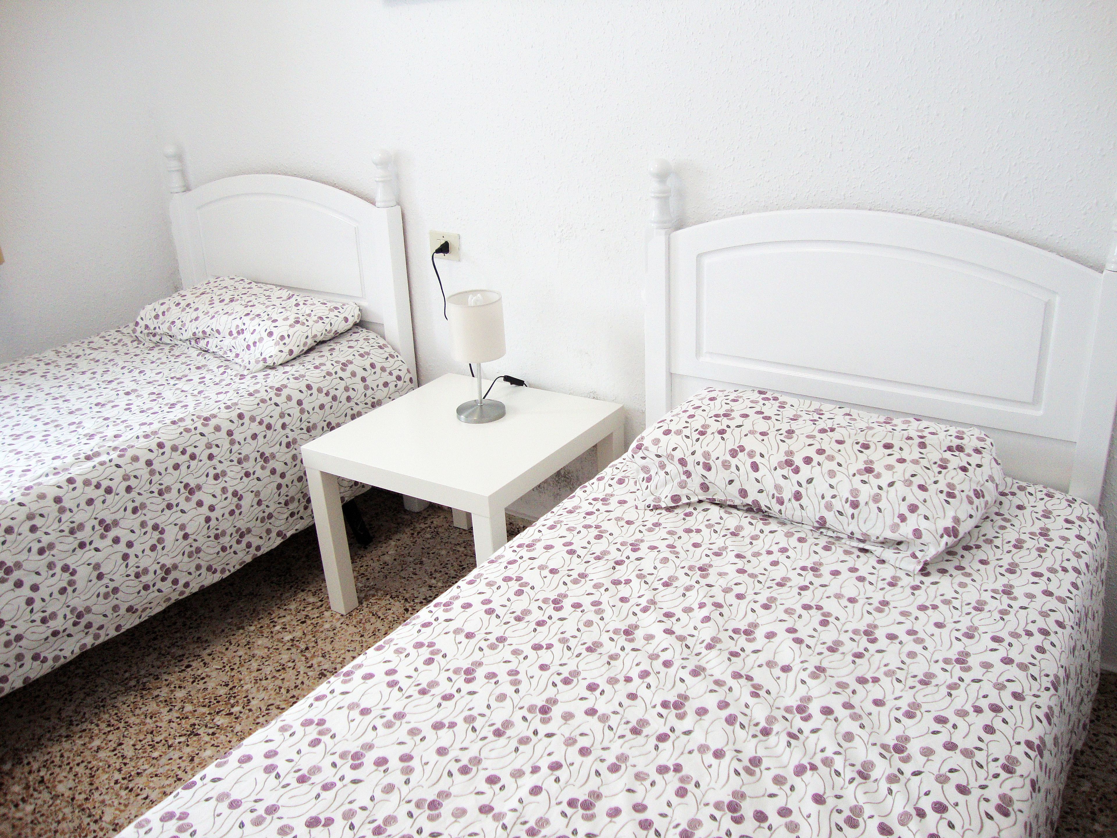 A room with two single beds, each with a floral-patterned bedspread, a small bedside table with a lamp, and a white wall.