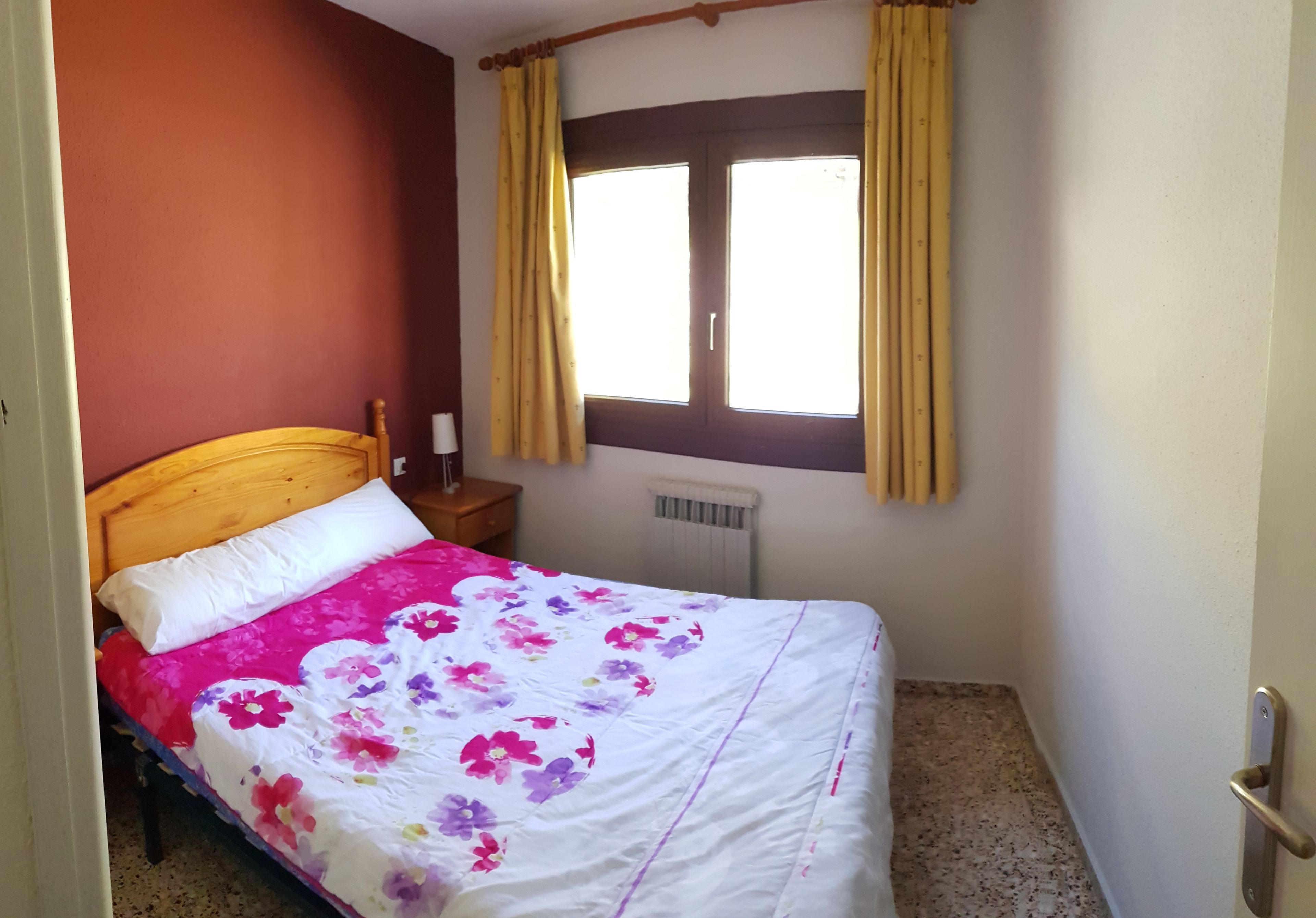A bedroom with a wooden bed, a floral bedspread, a window with curtains, a bedside table, and a radiator.