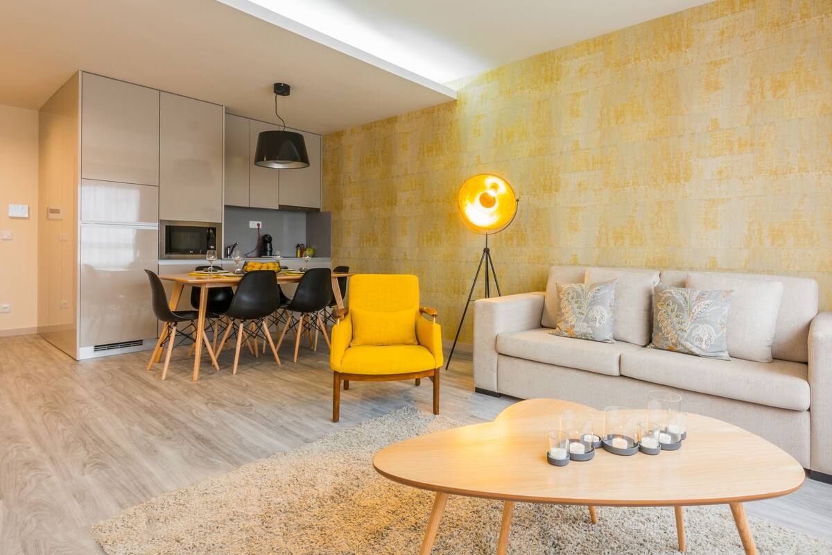 A modern living room with a sofa, a yellow chair, a dining table with chairs, a coffee table with candles, and a kitchen area with cabinets and a pendant light.