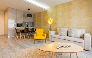 Uma sala de estar moderna com sofá, uma poltrona amarela, uma mesa de jantar com cadeiras, uma mesinha de centro com velas e uma área de cozinha com armários e uma luminária pendente.