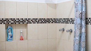 Un baño con baldosas color beige, un borde de mosaico blanco y negro, una cortina de ducha con estampado azul y blanco, y dos botellas de jabón Vanart.