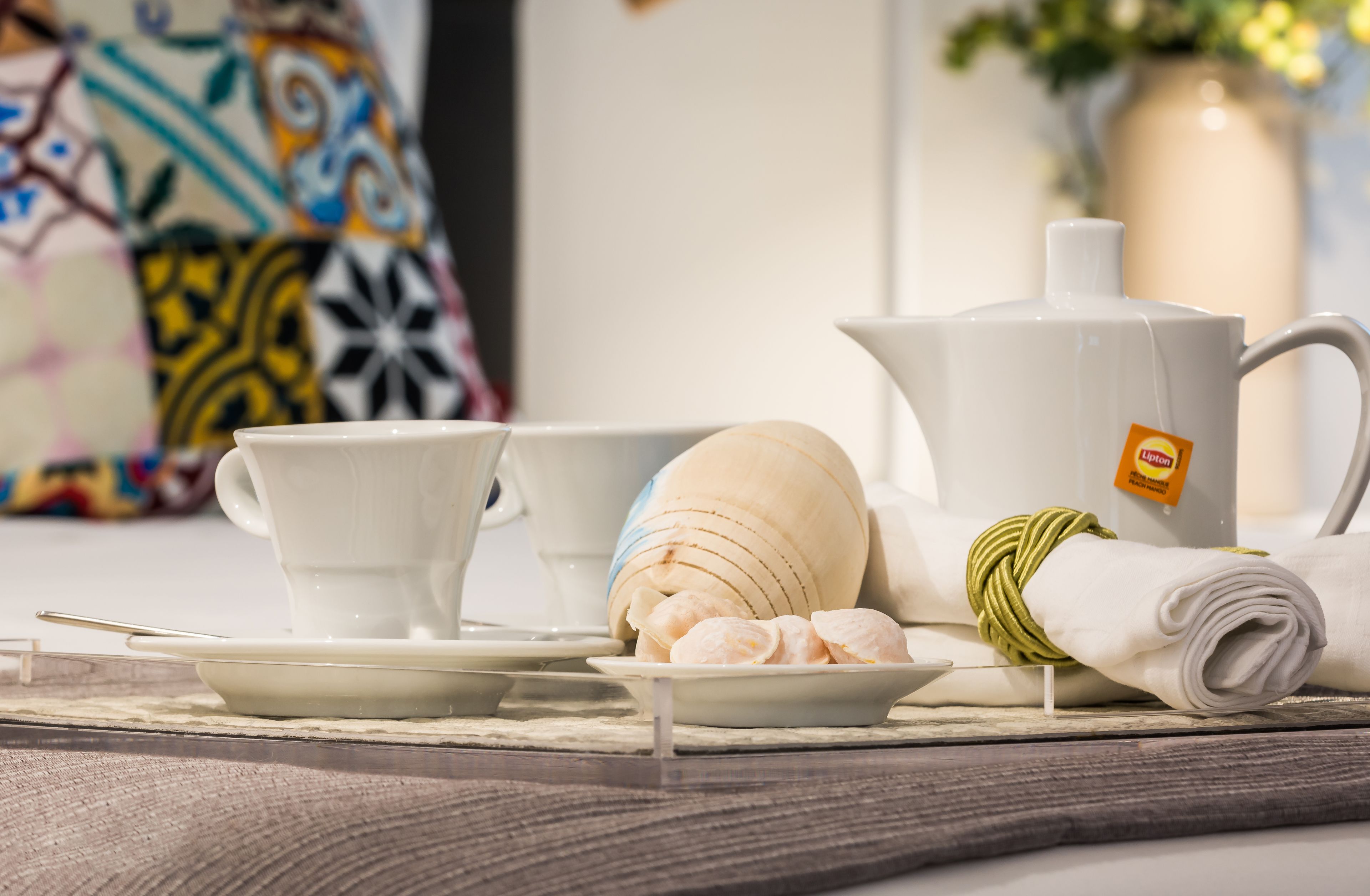 A tea set with a teapot, cups, and saucers on a table, with a rolled towel and a decorative vase in the background.
