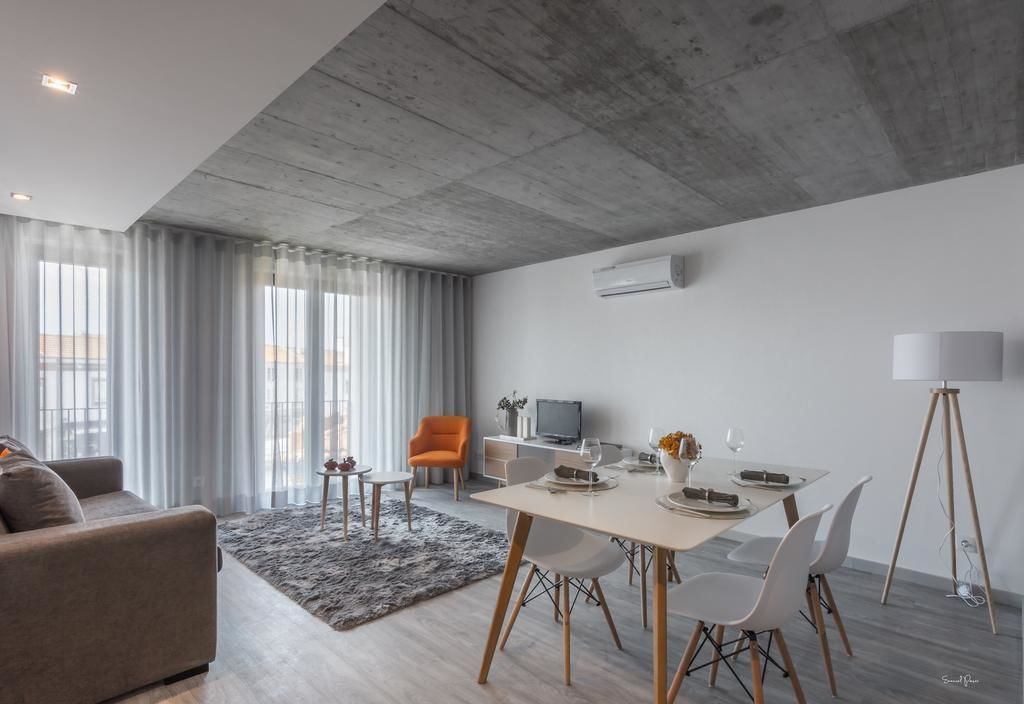 Una sala de estar moderna con comedor, cortinas grises, un sofá gris, un sillón pequeño naranja y una lámpara de pie blanca.