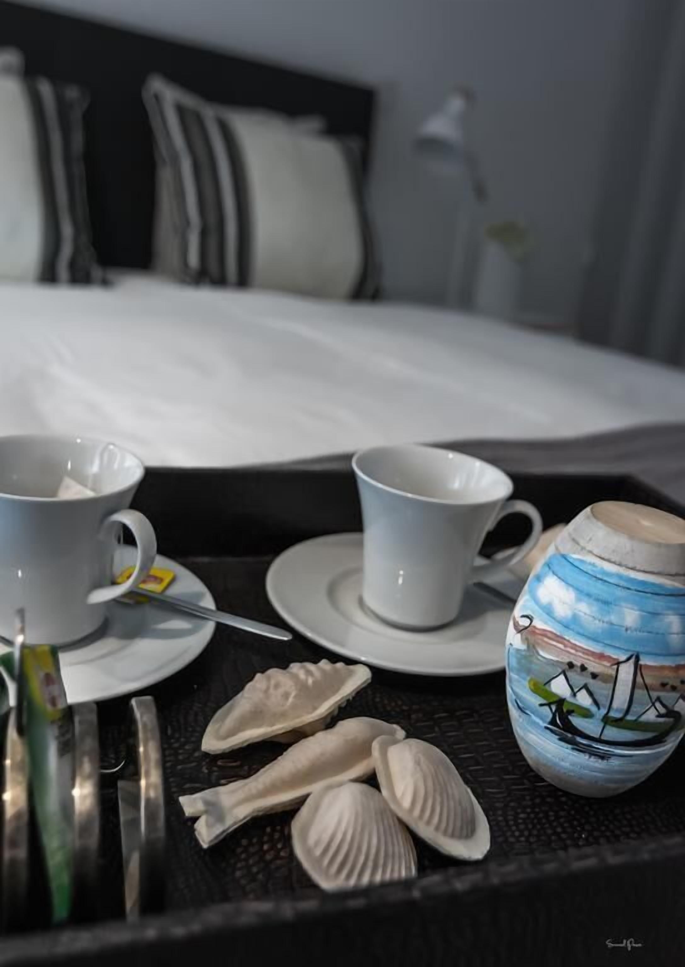 A tray with two white cups, a teapot, and seashells on a bed with a black headboard and white bedding.