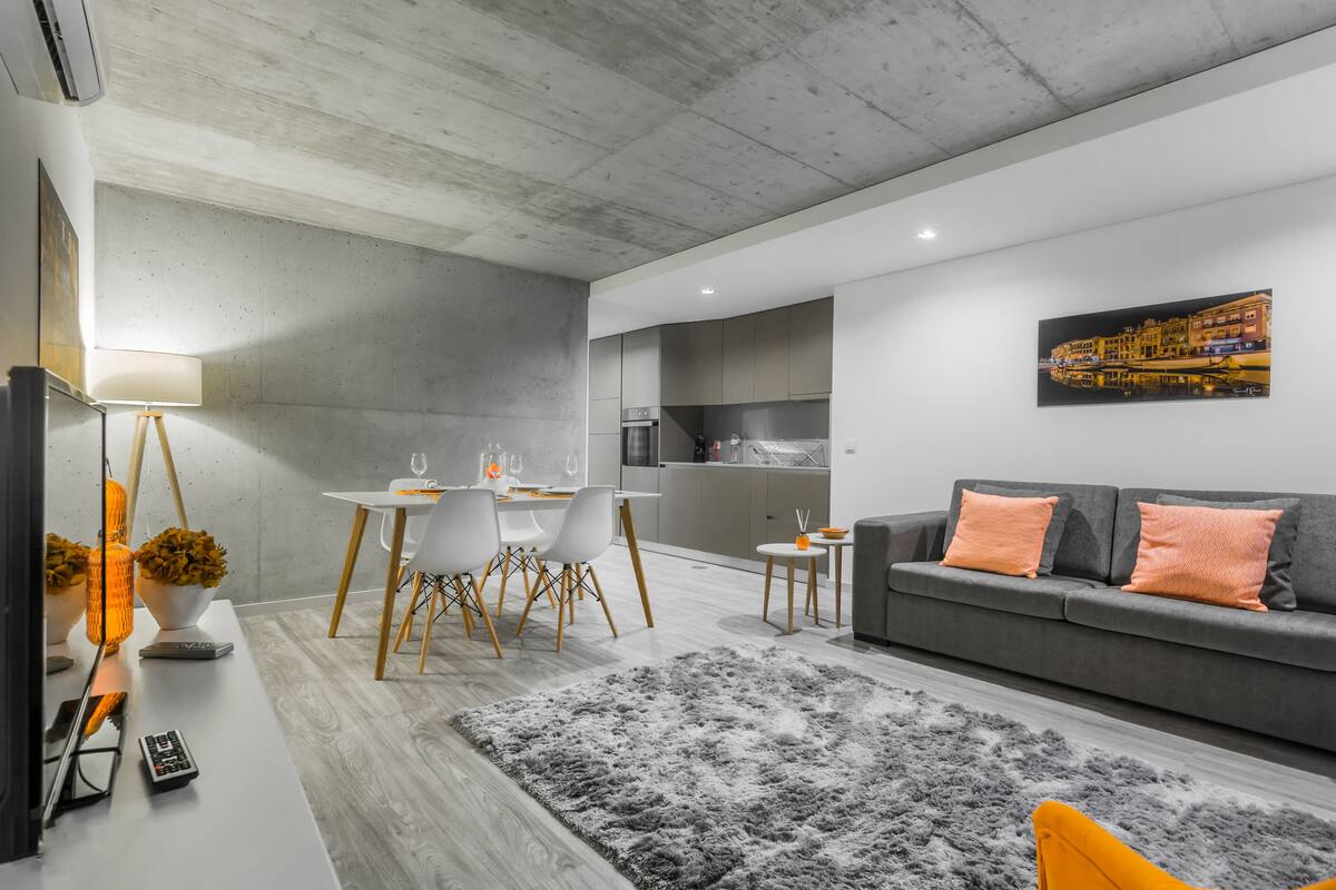 A modern living room with a grey sofa, a dining area with white chairs, and a kitchen in the background.