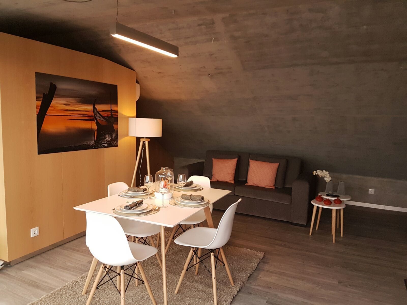 Un área de comedor con una mesa dispuesta para cuatro personas, un sofá, una lámpara y una pequeña mesita con un jarrón.