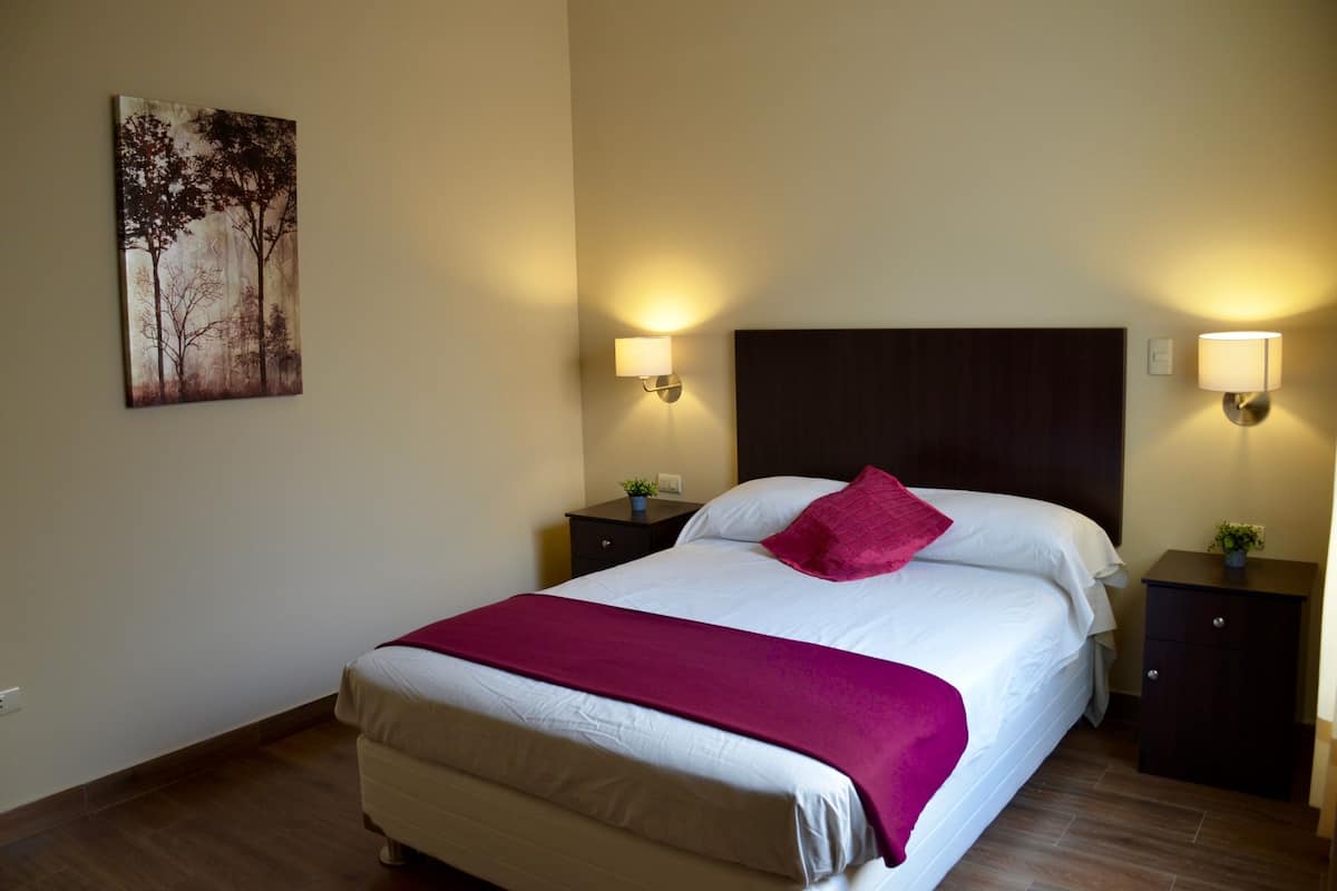 Una cama bien hecha con edredón blanco y una manta rosa, flanqueada por dos mesitas de noche con lámparas, en una habitación con suelo de madera y un cuadro colgado en la pared.