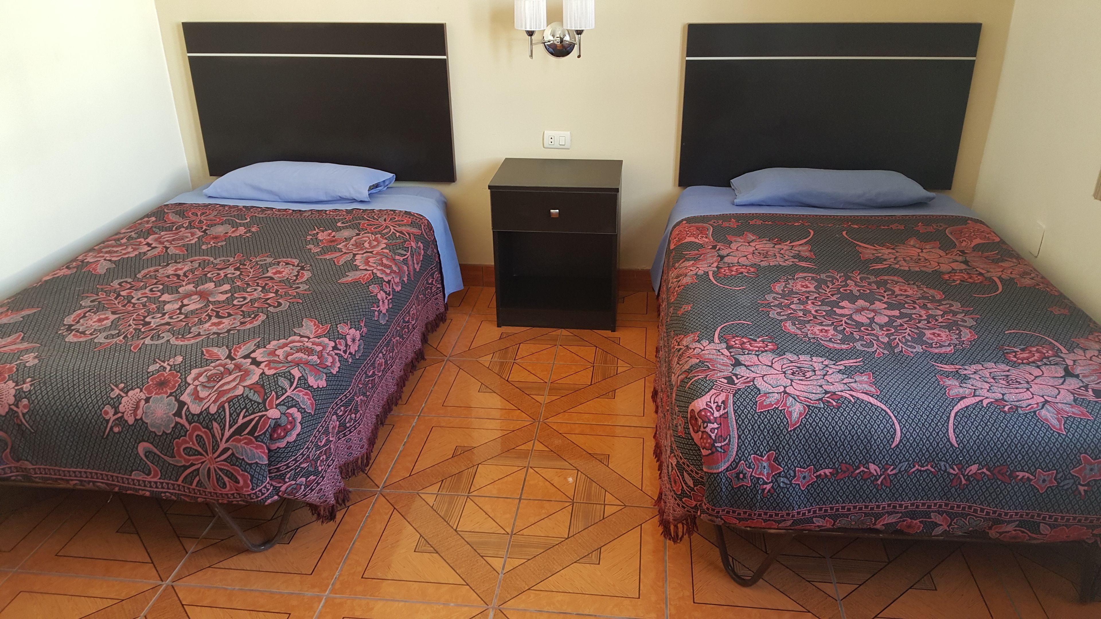 Quarto com duas camas, cada uma com colcha estampada e almofada azul. Entre as camas, um pequeno criado-mudo preto com um interruptor de luz.