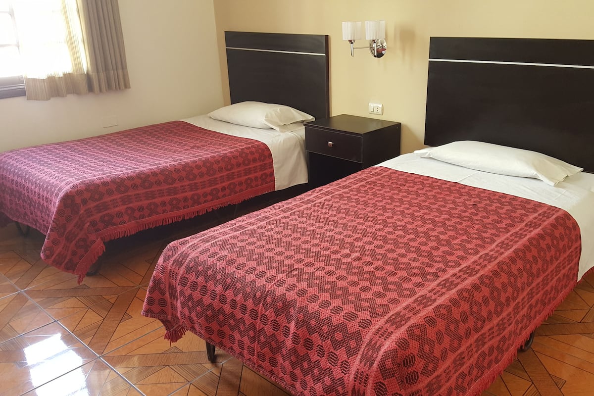 Dos camas individuales con cubrecamas rojos estampados, una mesita de noche entre ellas y una lámpara de pared.
