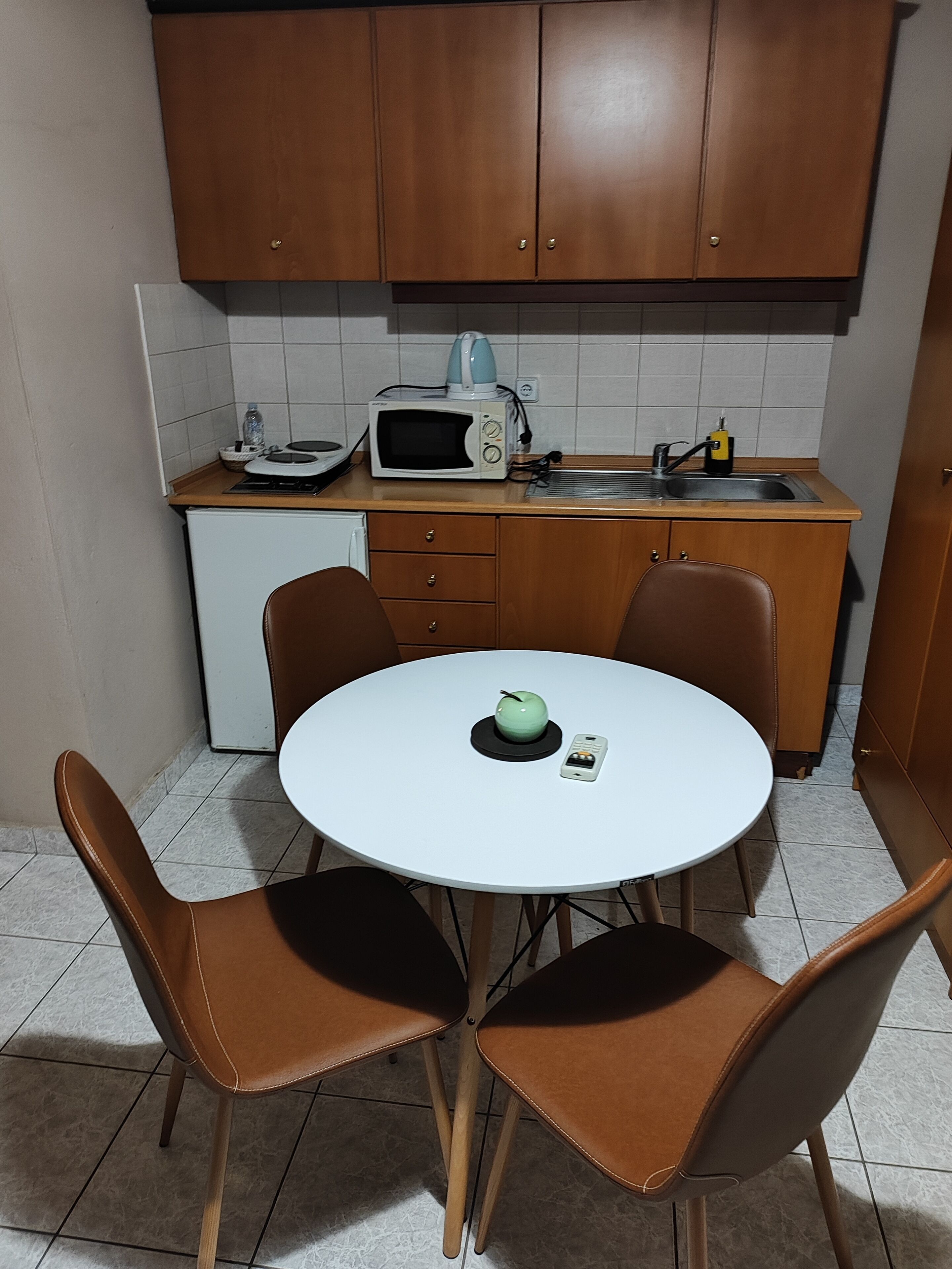 Une petite cuisine équipée d’un four à micro-ondes, d’une bouilloire et d’un évier. On y trouve une table ronde avec des chaises, et une pomme verte posée dessus.
