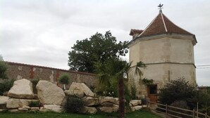 A stone building with a red-tiled roof, surrounded by a wooden fence and a grassy area with large rocks and a palm tree. - Le Colombier de la Graverie (Senots)