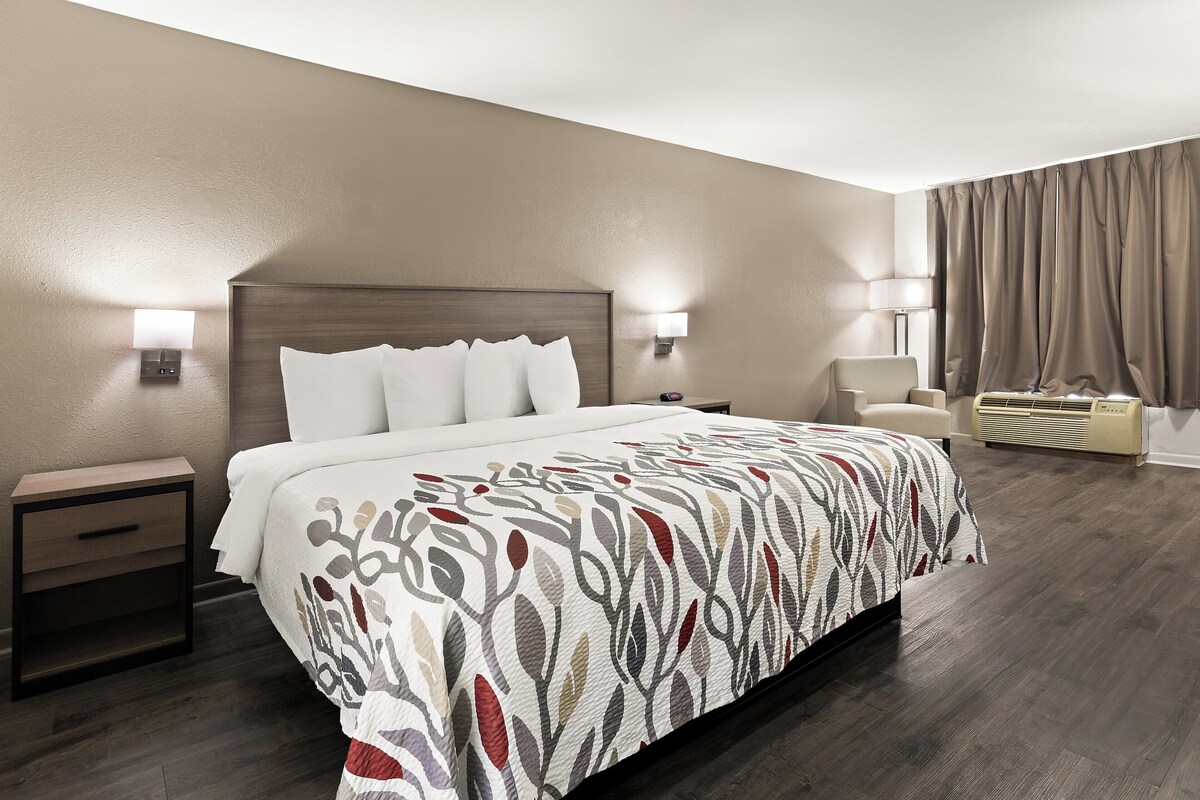 A hotel room with a large bed, bedside tables, a chair, an air conditioning unit, and a lamp.