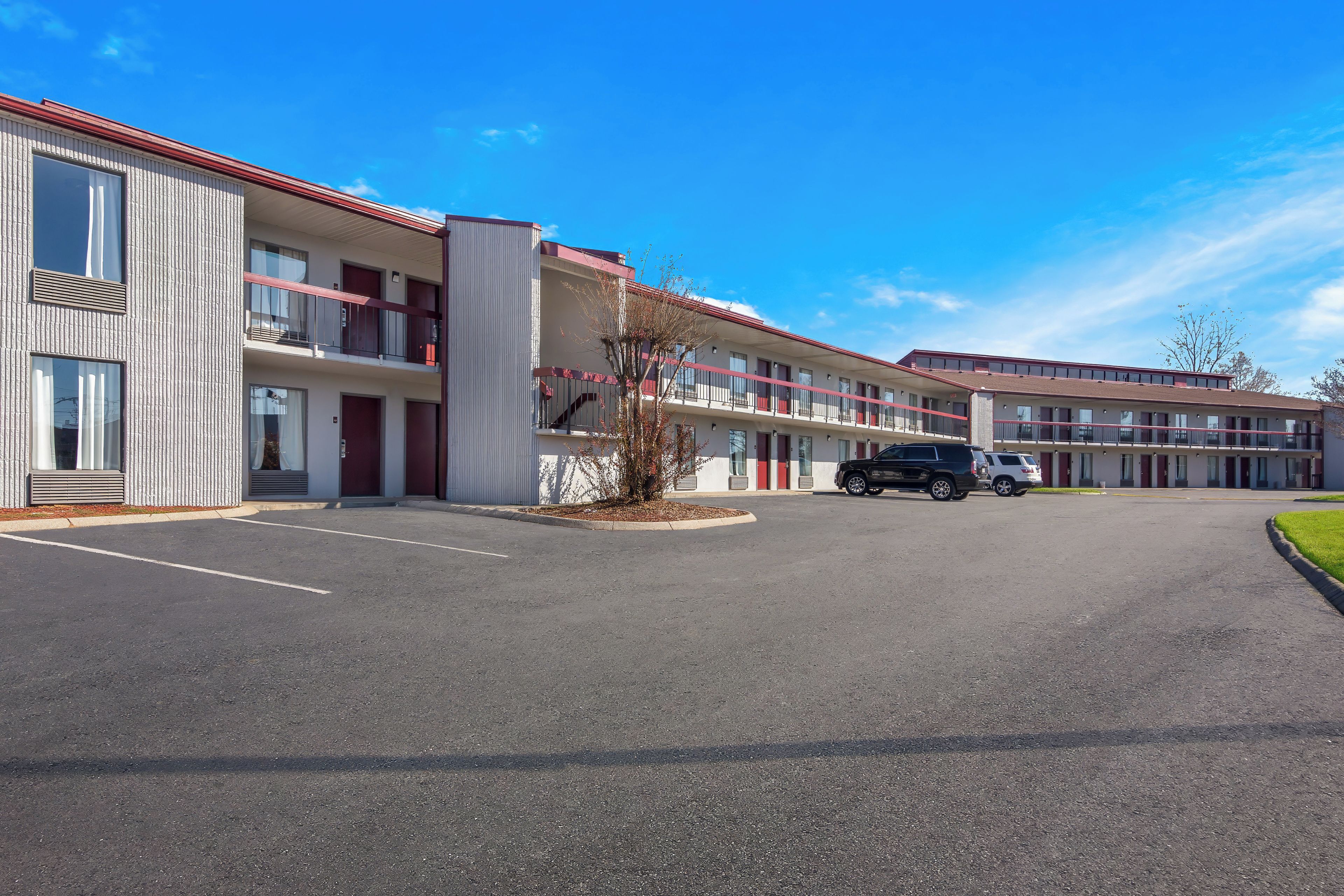 A multi-story motel with a parking lot, featuring a row of rooms with balconies, a central courtyard, and a clear blue sky.