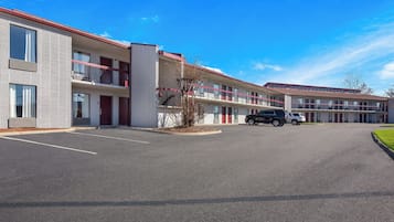 A multi-story motel with a parking lot, featuring a row of rooms with balconies, a central courtyard, and a clear blue sky.