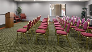 A conference room with rows of red chairs and a podium, carpeted floor, and ceiling lights. - Red Roof Inn & Suites Monroe, NC (Monroe)