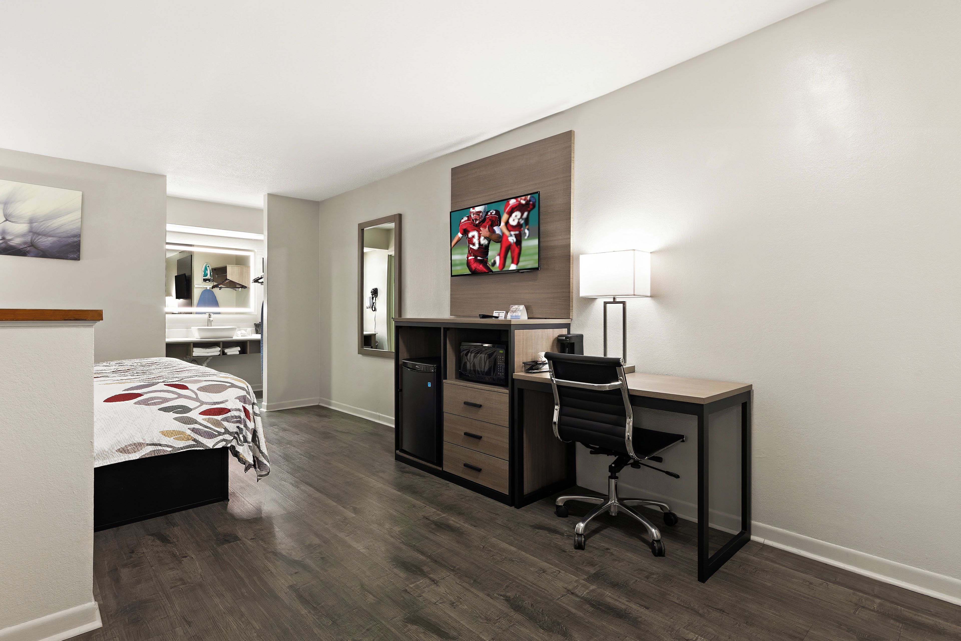 A hotel room with a bed, a desk, a chair, a mirror, a TV, and a lamp.