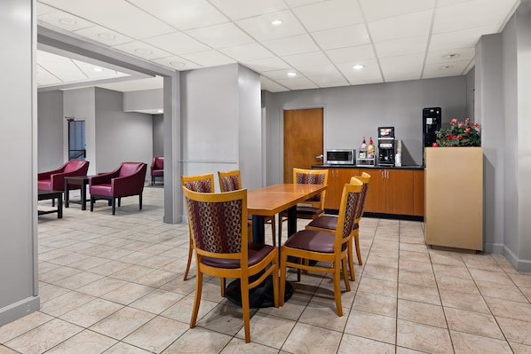 A hotel dining area with a wooden table and chairs, a kitchenette, and a seating area with red chairs.
