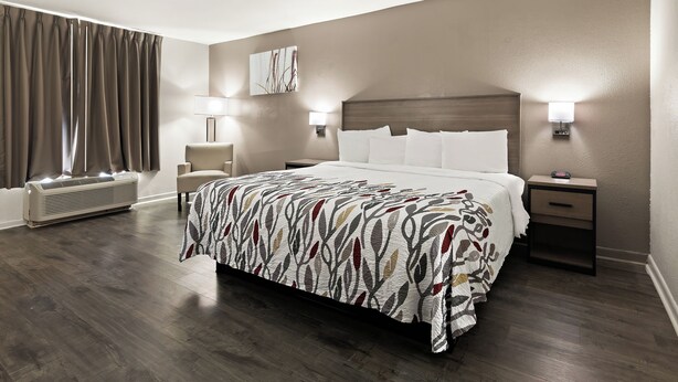 A hotel room with a large bed, two bedside tables with lamps, a wall-mounted heater, and a window with curtains. - Red Roof Inn & Suites Monroe, NC (Monroe)
