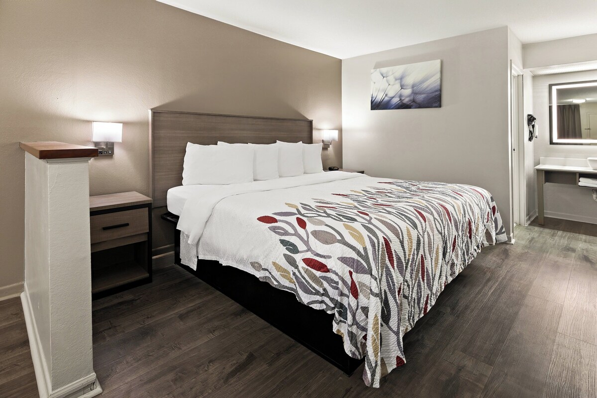 A hotel room with a large bed, bedside tables, a nightstand, a wall-mounted light, a painting, and a bathroom with a mirror and sink.
