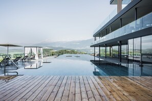 Piscina infinita moderna con cubierta de madera, sillones de descanso y una terraza acristalada con vistas a un paisaje montañoso.