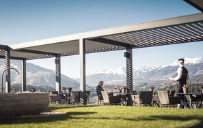 Un espacio al aire libre con mesas y sillas, una estructura de pérgola y montañas al fondo.
