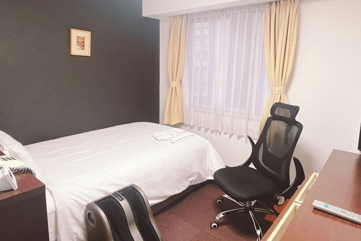 Habitació d'hotel amb llit, escriptori, cadira, telèfon i finestra amb cortines.