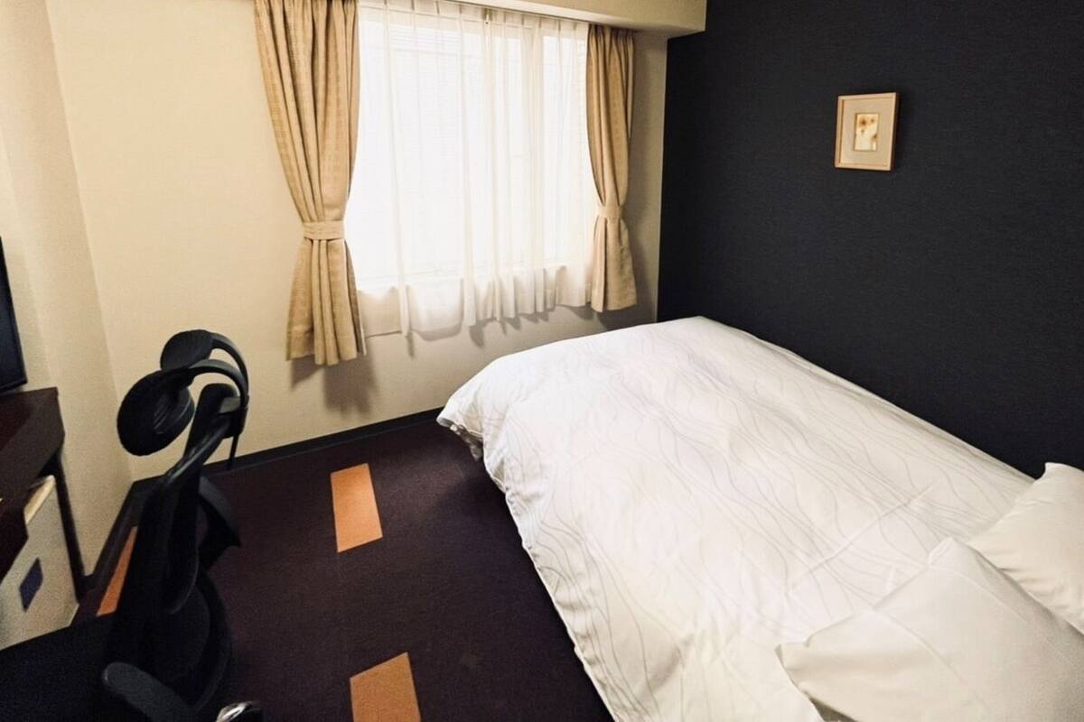 A hotel room with a bed, a desk, a chair, a window with curtains, and a wall-mounted picture.