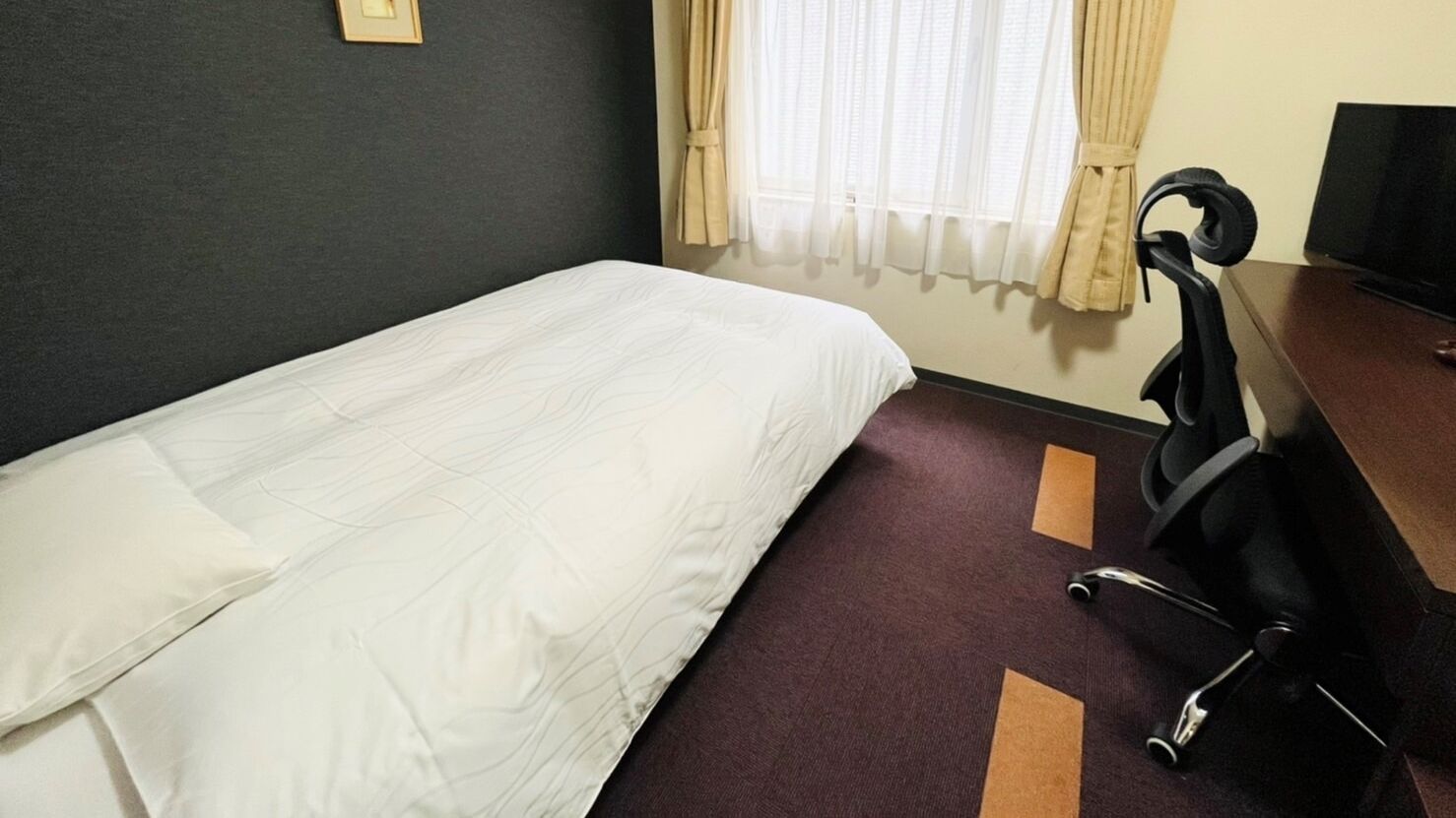 A hotel room with a single bed, a desk with a chair, a television, and a window with curtains.