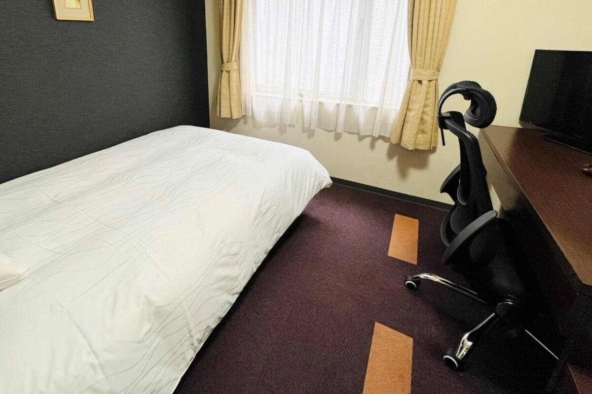 A hotel room with a single bed, a desk with a chair, a television, and a window with curtains.