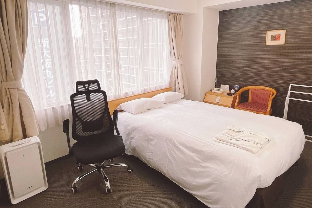 A hotel room with a bed, a desk, a chair, a radiator, and a window with curtains.