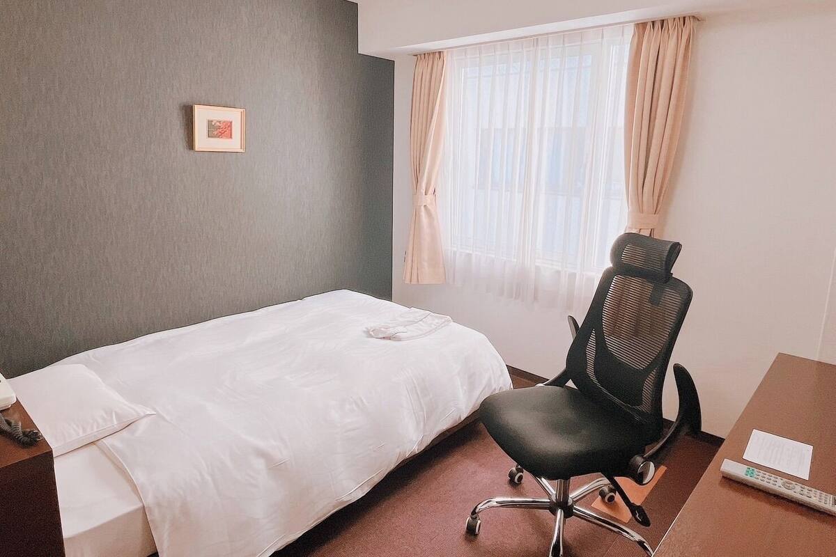 A hotel room with a single bed, a desk with a chair, a telephone, and a window with curtains.