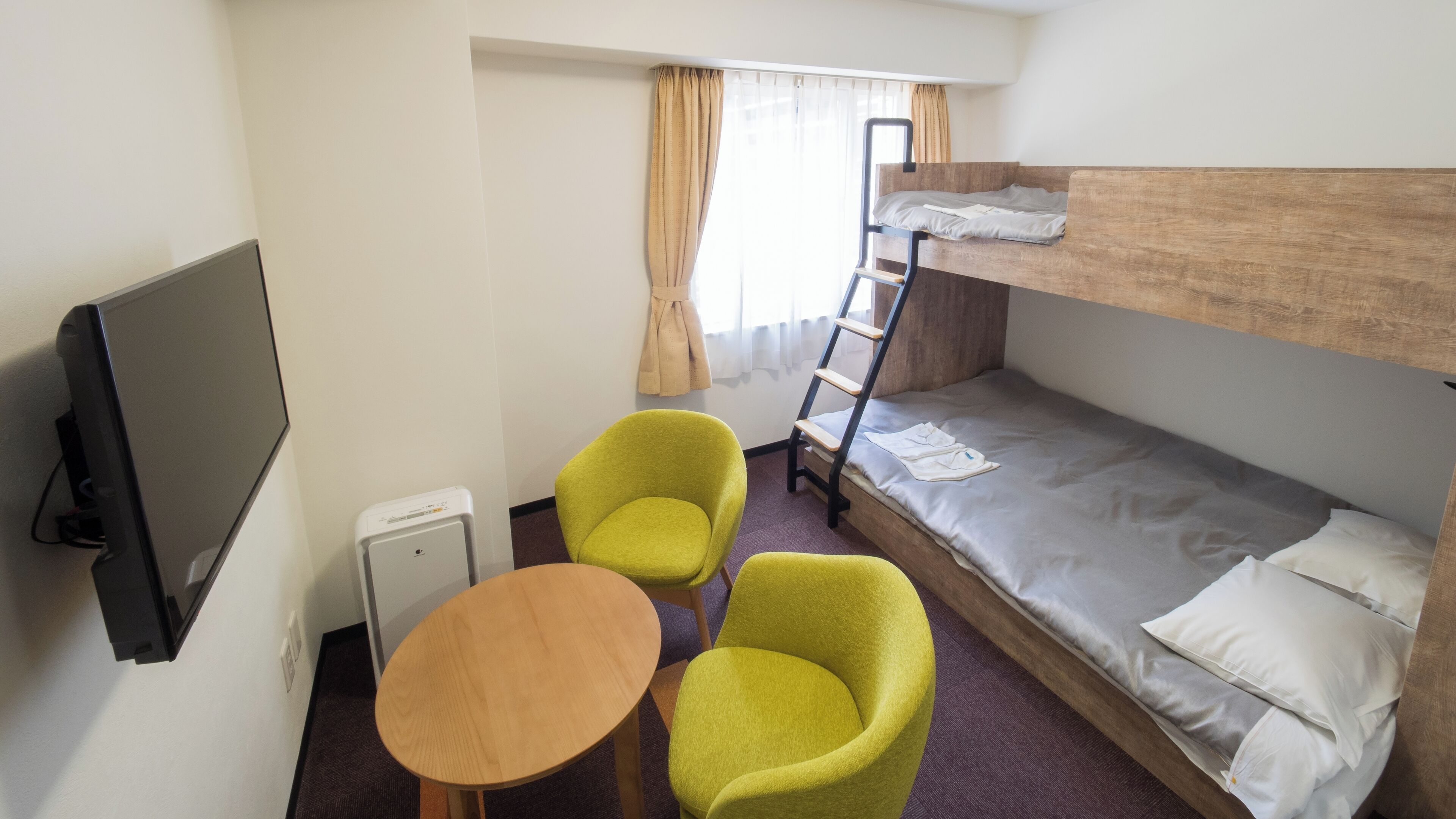 A room with a bunk bed, a television, two chairs, a small table, and a window with curtains.