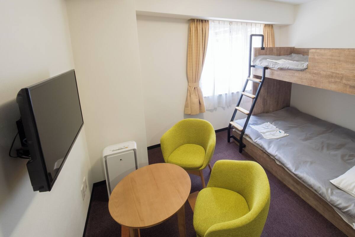 A room with a bunk bed, a television, two chairs, a small table, and a window with curtains.