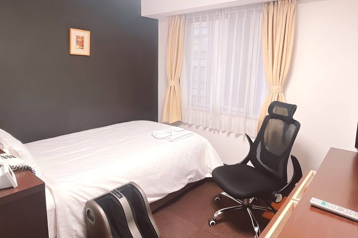 A hotel room with a bed, a desk with a chair, a telephone, and a window with curtains.