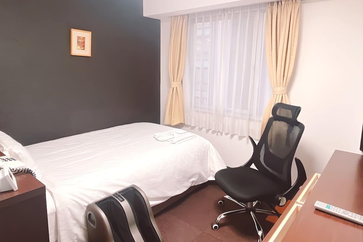 Habitació d'hotel amb un llit, una taula de nit amb cadira, un telèfon i una finestra amb cortines.