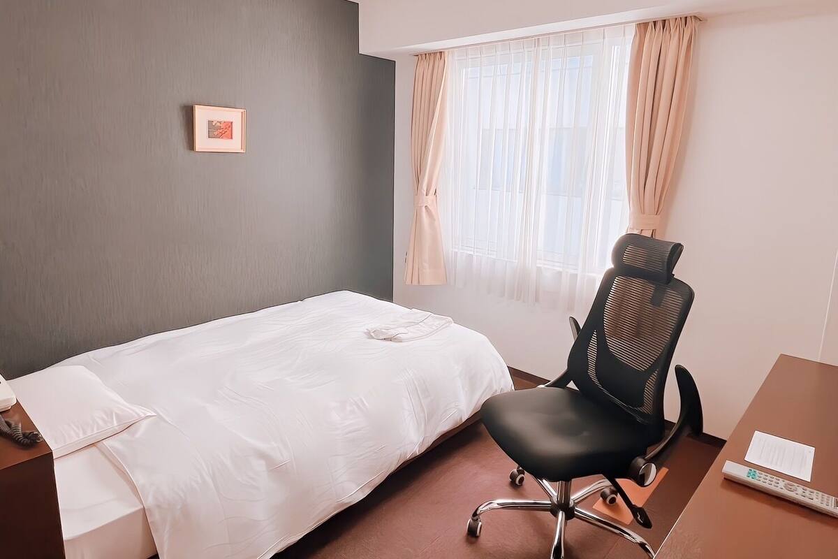 A hotel room with a single bed, a desk with a chair, a telephone, and a window with curtains.