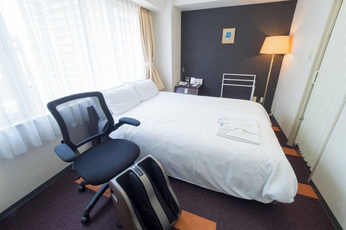 A hotel room with a bed, a desk, a chair, a lamp, and a small picture on the wall.