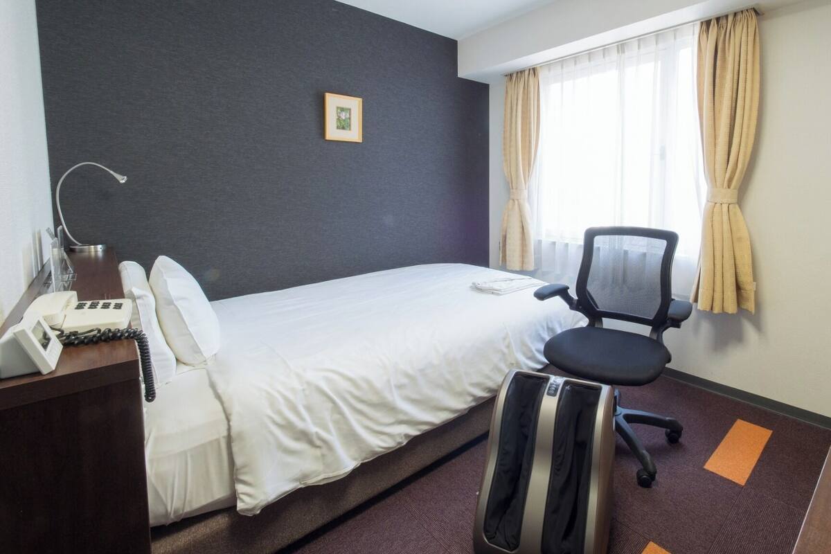 A hotel room with a bed, a desk with a telephone, a chair, a television, and a window with curtains.