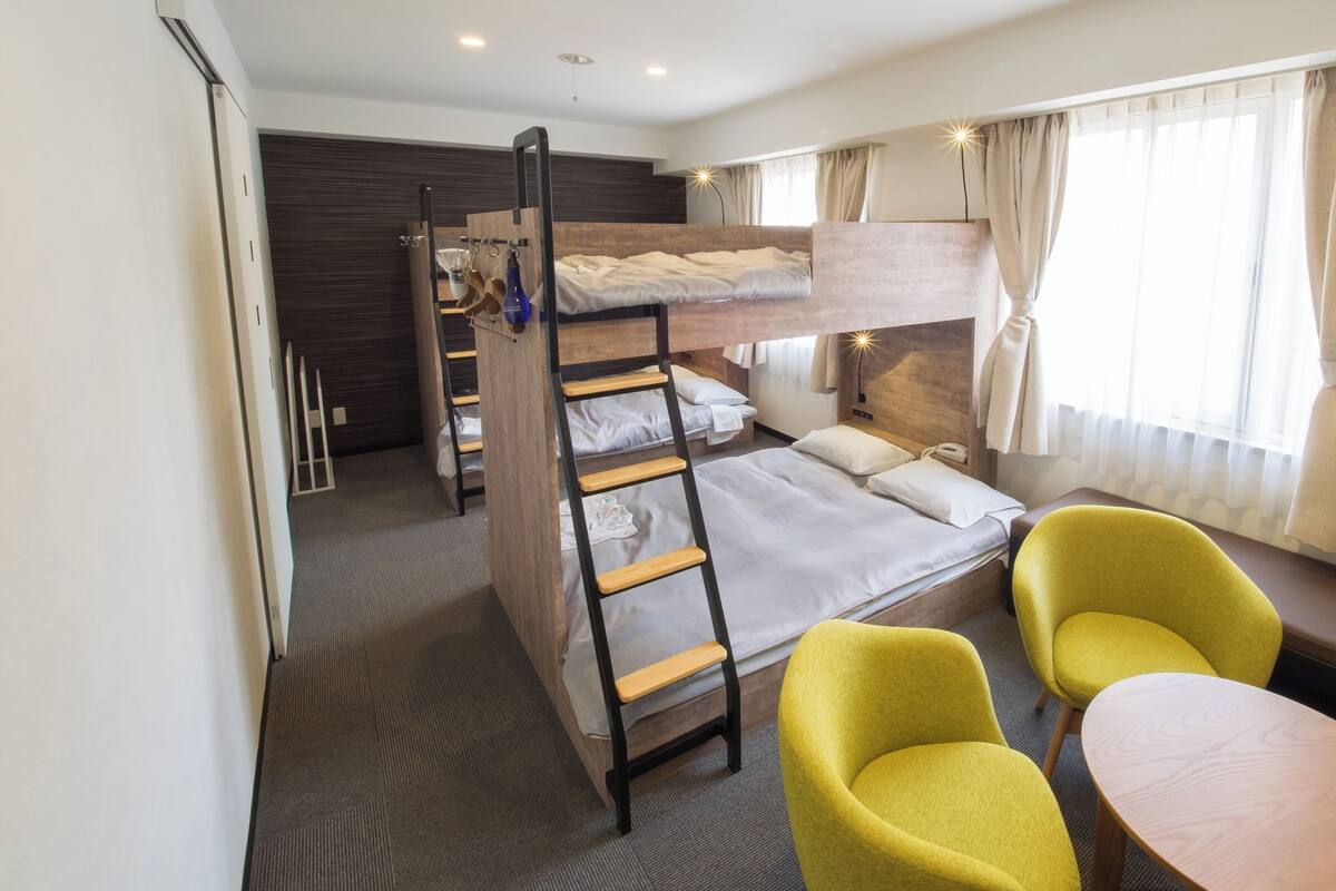 A bunk bed room with a ladder, a small table, two chairs, and a window with curtains.