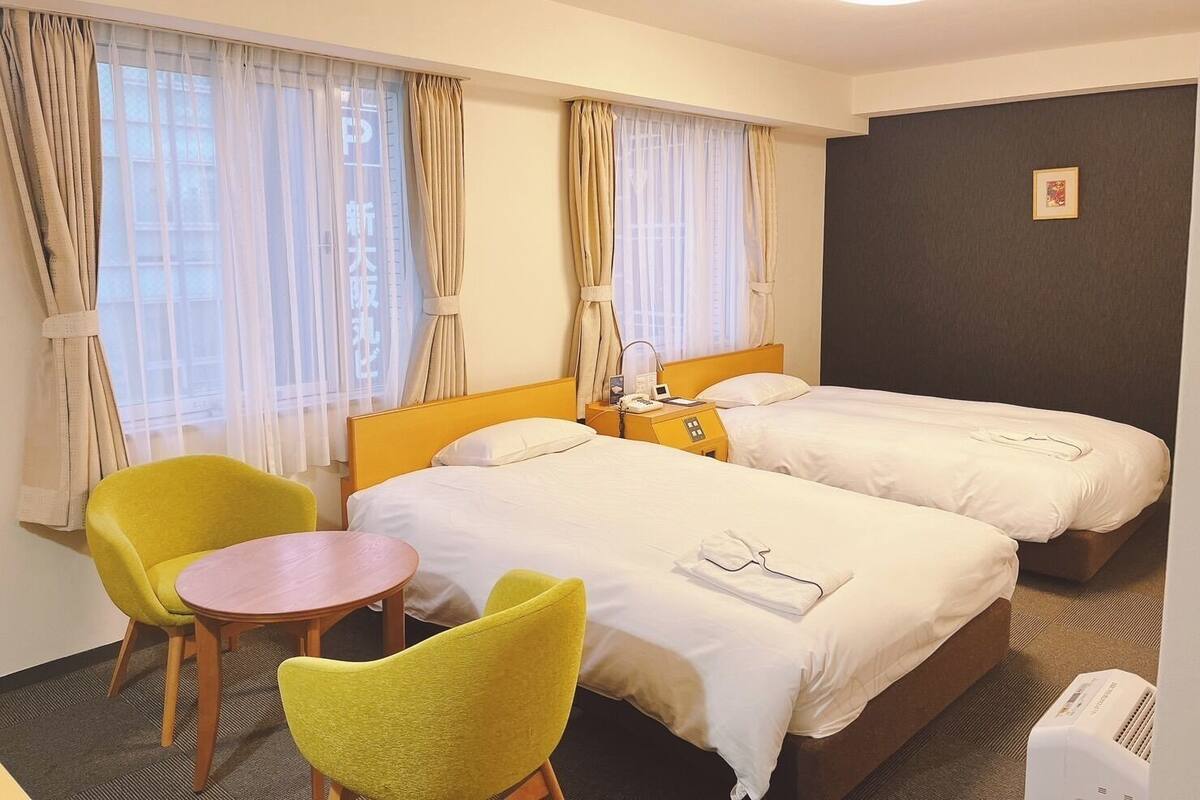 A hotel room with two beds, a small table, two chairs, a bedside table with a lamp, and a wall-mounted air conditioner.