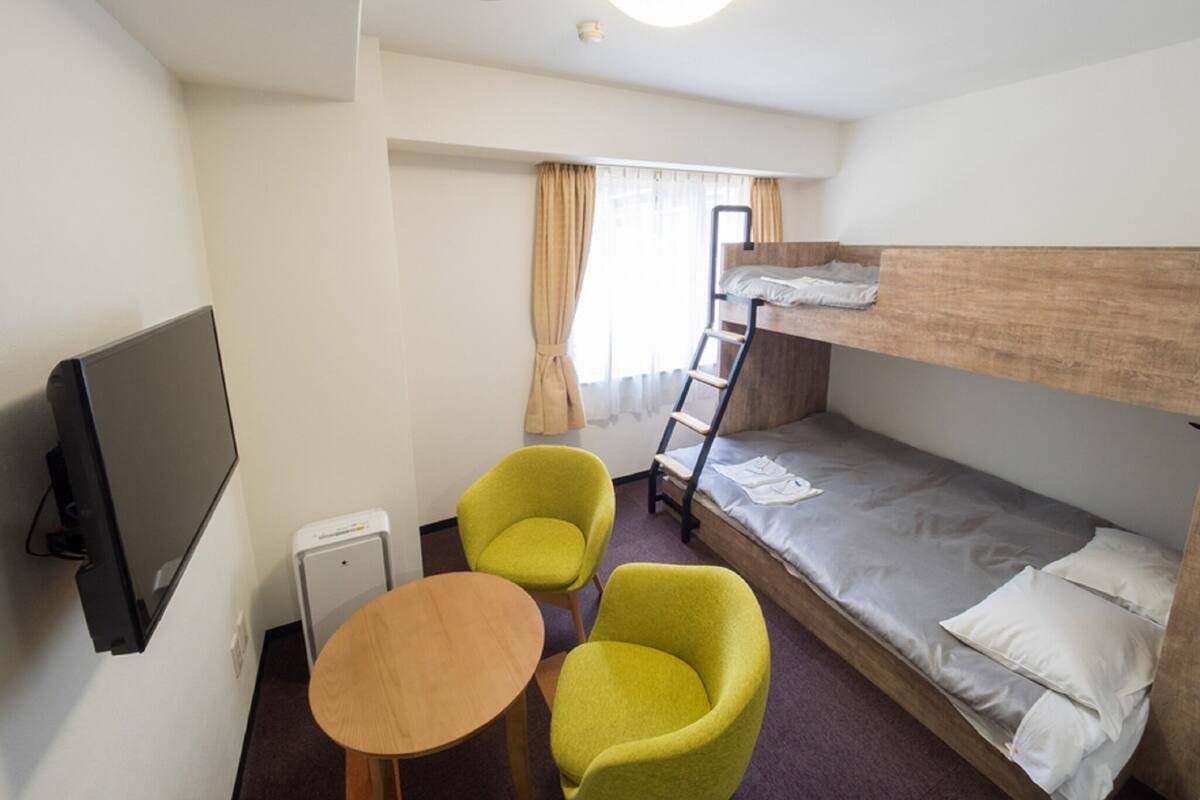 A hotel room with a bunk bed, a television, a small table, two chairs, and a window with curtains.