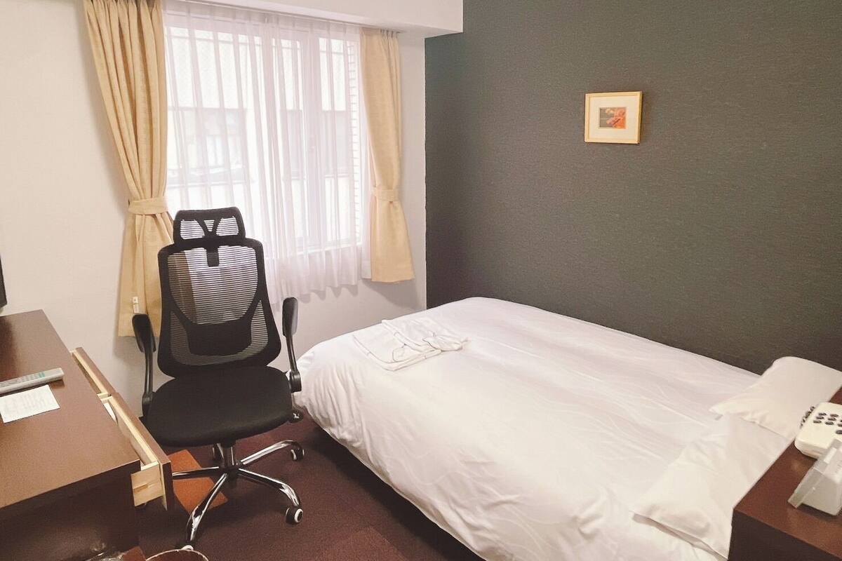 A hotel room with a bed, a desk with a chair, a telephone, and a window with curtains.