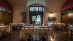 A dining area with tables set for four, wooden chairs, and a large window with curtains. - Casale La Sterpaia (San Giuliano Terme)