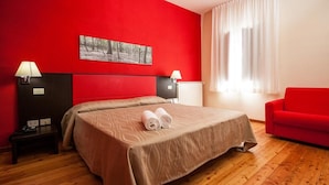 A hotel room with a red accent wall, a bed with a beige bedspread, two bedside lamps, a red sofa, and a window with white curtains. - Casale La Sterpaia (San Giuliano Terme)
