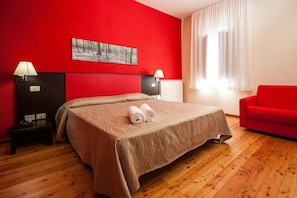 A hotel room with a red accent wall, a bed with a beige bedspread, two bedside lamps, a red sofa, and a window with white curtains. - Casale La Sterpaia (San Giuliano Terme)